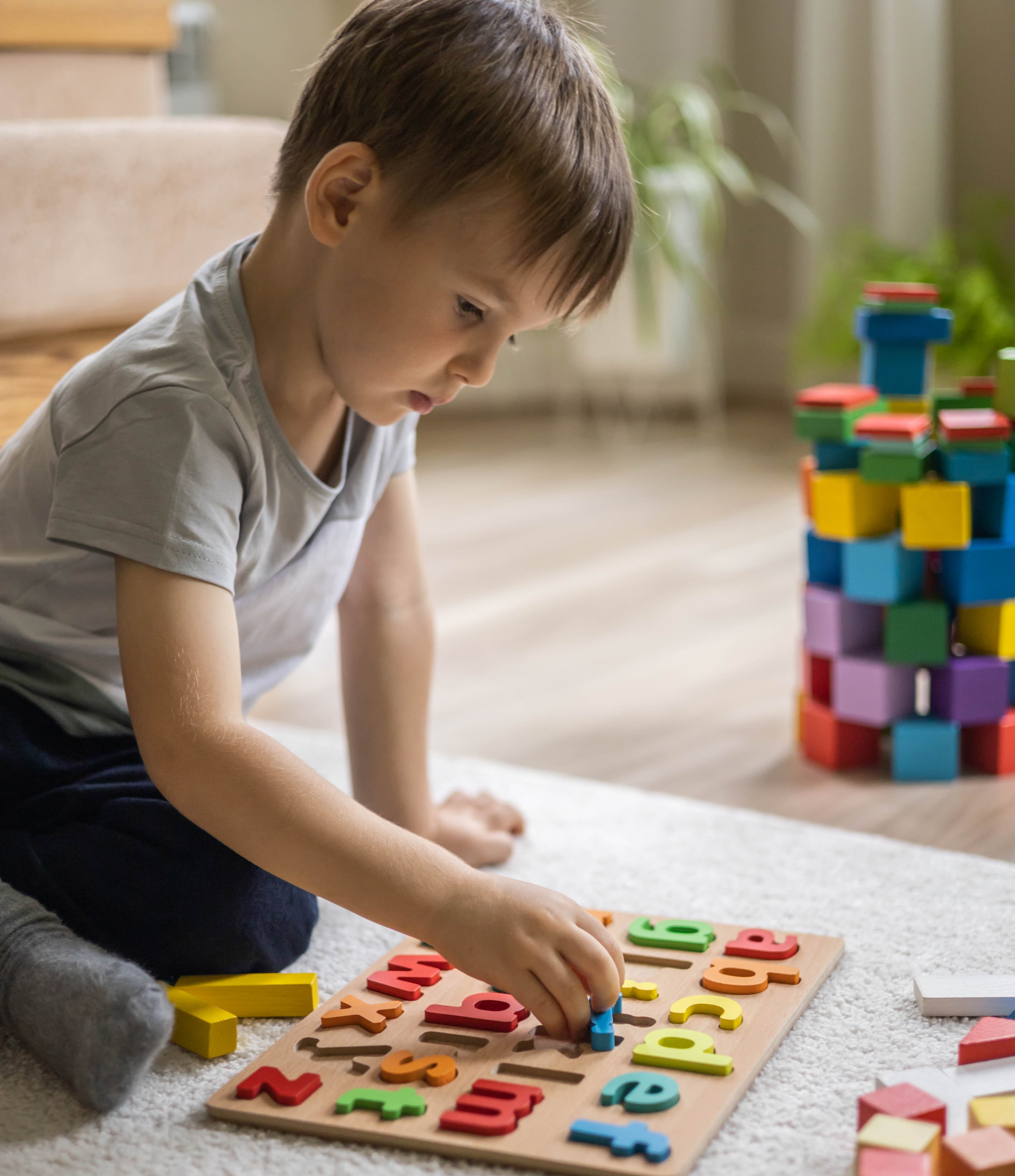 A young child kneeling on the living-room floor, concentrating on a wooden alphabet puzzle in warm afternoon light.