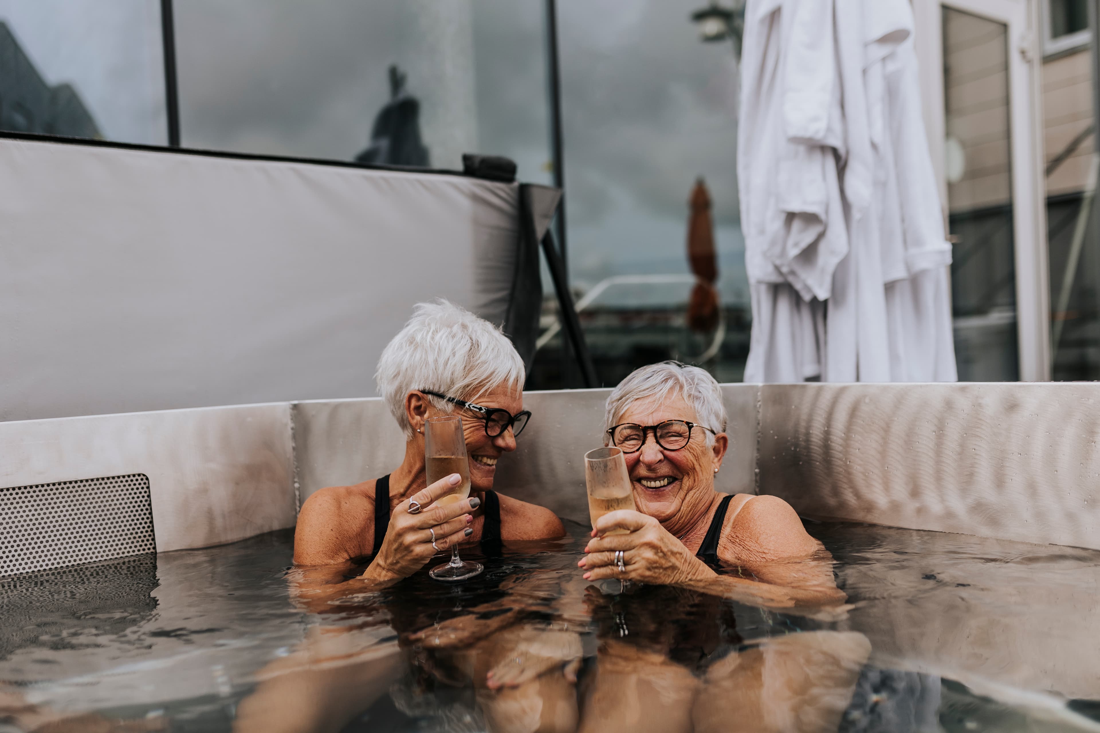 Two older women laughing together in a hot tub holding wine glasses, a moody overcast sky reflected in the window behind them.