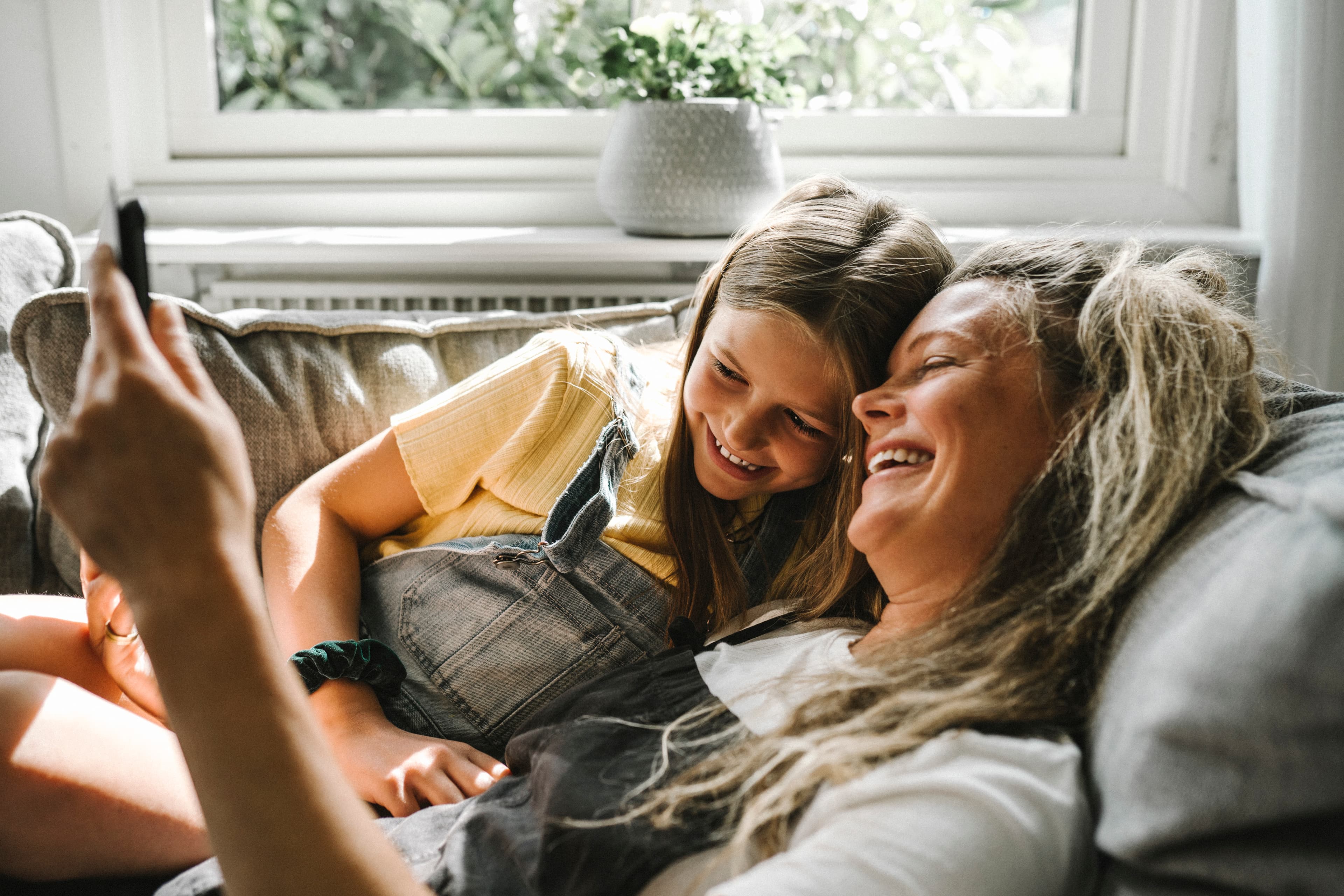 A mother and daughter laughing together on a couch, looking at a phone in a sunlit living room.