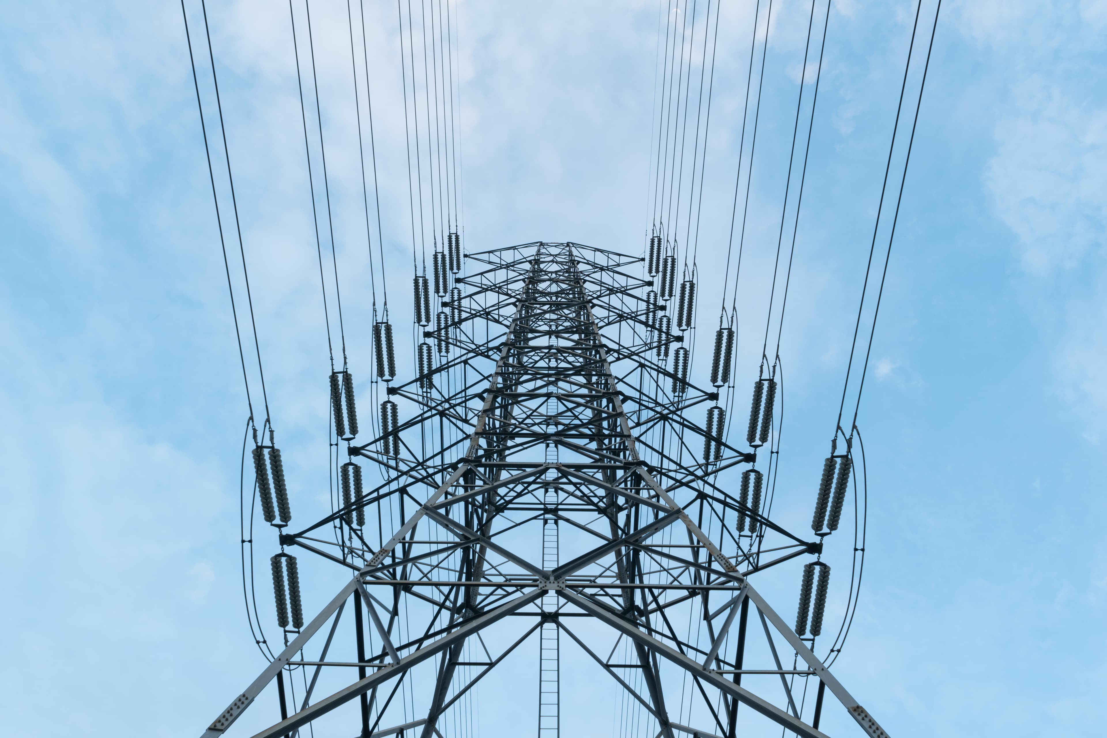 A residential power pylon rising against a clear blue sky — the lines-company asset homeowners are typically quoted thousands to upgrade.