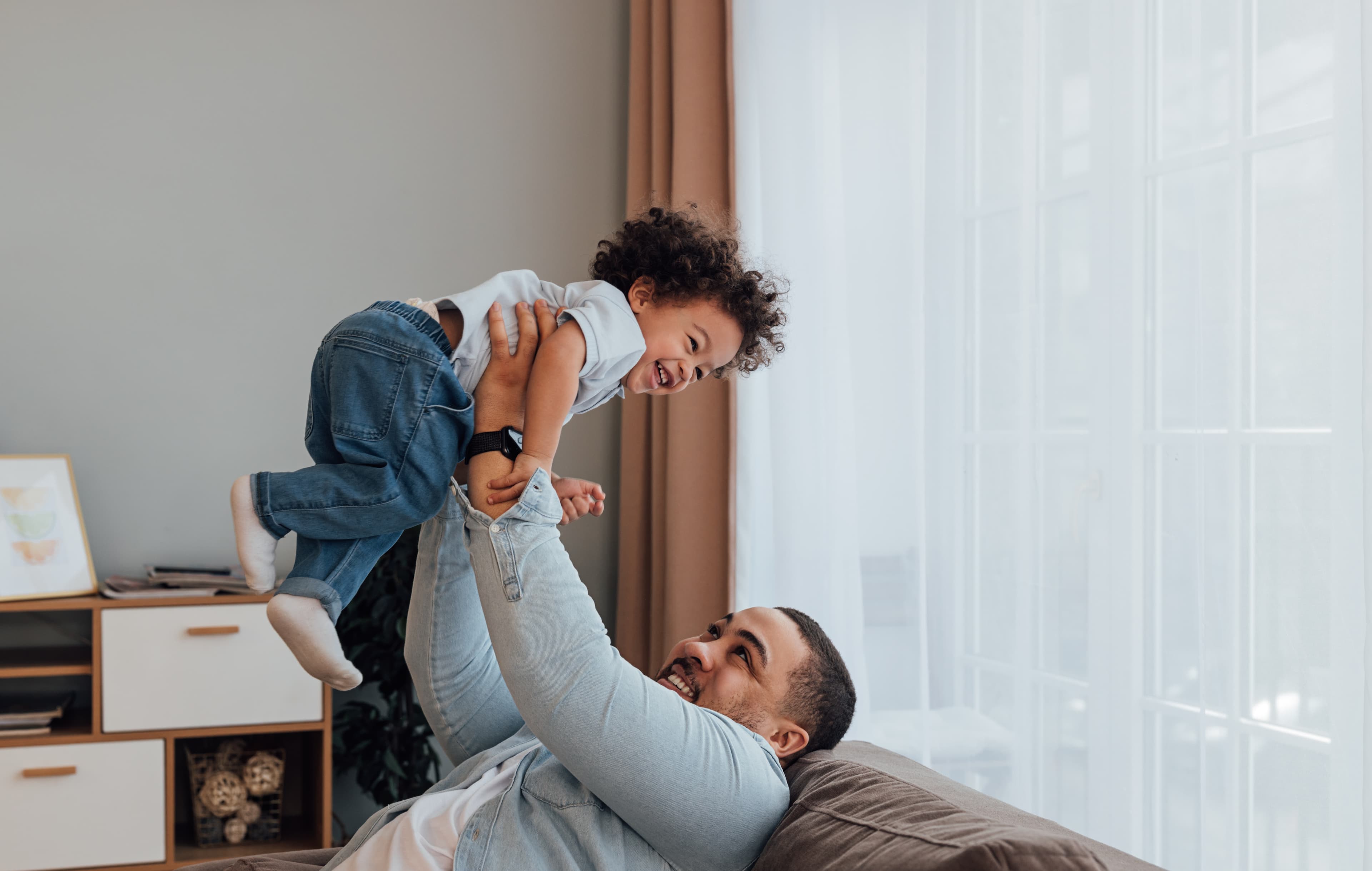 A father lifting his laughing child into the air in a sunlit living room